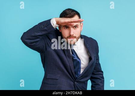 Far future. Portrait of handsome man wearing official style suit looking into distance, holding hand over eyes and watching horizon with attentive view. Indoor studio shot isolated on blue background. Stock Photo