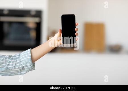 Female hand showing smartphone with blank screen, standing in kitchen interior, mockup Stock Photo