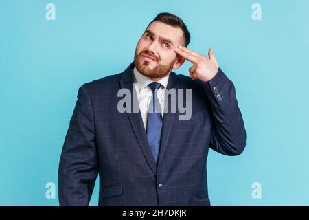 Depression, suicide gesture. Portrait of businessman wearing official style suit with finger gun pointed to head, shooting himself with hand pistol. Indoor studio shot isolated on blue background. Stock Photo