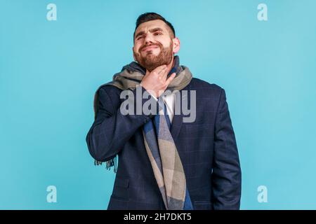 Young adult man with beard wearing official style suit, being wrapped in scarf, touching his neck, suffering from sore throat and high temperature. Indoor studio shot isolated on blue background. Stock Photo