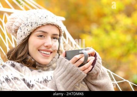 Happy woman holding coffee cup looks away in autumn on hammock in a forest Stock Photo