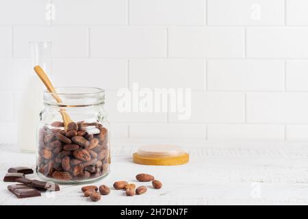 Cocoa beans in a glass jar on a light kitchen table. Culinary food cooking background for making traditional chocolate. Selective focus Stock Photo