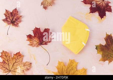 Yellow respirator KN95 or FFP2 mask on pink background, autumn maple leaves, copy space. Fall and new rules against coronavirus Covid-19 concept Stock Photo