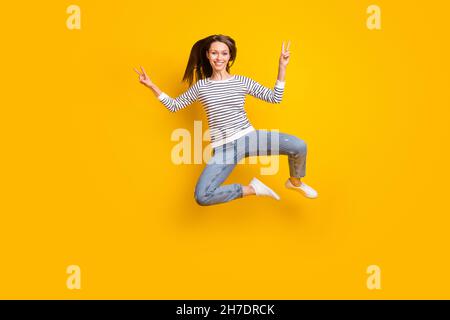 Full size photo of lovely brunette millennial lady jump show v-sign wear sweater jeans sneakers isolated on yellow color background Stock Photo