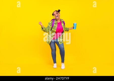 Full body photo of cheerful afro american woman wear backpack hold drink winner isolated on yellow color background Stock Photo