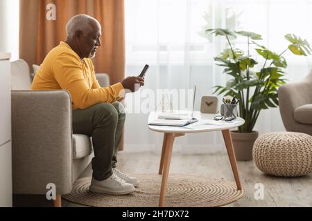 Mature Black Man Using Smartphone Texting Sitting At Laptop Indoor Stock Photo