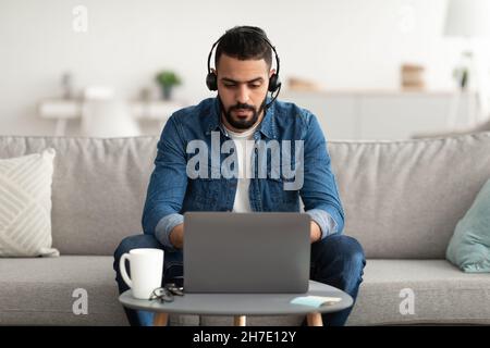 Young serious Arab man wearing headphones, using laptop computer, working online from home Stock Photo