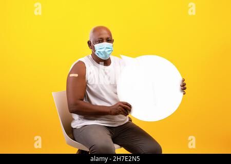 Vaccinated senior black guy with band aid plaster after anti covid vaccine shot holding empty speech bubble with mockup Stock Photo