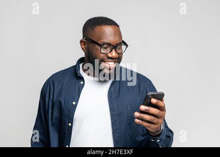 Portrait of cheerful young black guy sending message on mobile phone over white background. Happy African-American man using smartphone, enjoying chatting online, messaging in social networks Stock Photo