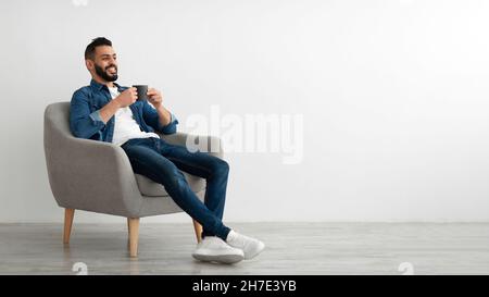 Full length of young Arab man drinking hot coffee in armchair against white studio wall, banner with free space Stock Photo