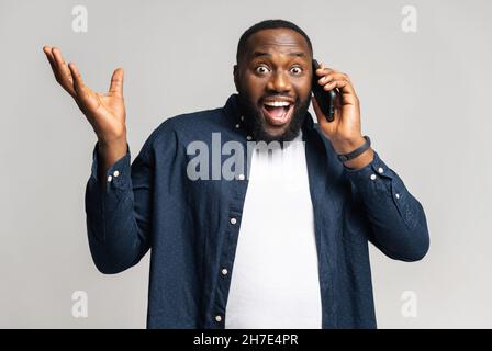 Wondered amazed happy African-American man in casual wear stands isolated on white, talking on smartphone, male student looking at camera with surprised face, spread hands during phone conversation Stock Photo