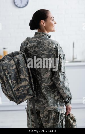 Side view of smiling soldier with backpack holding cap at home Stock Photo