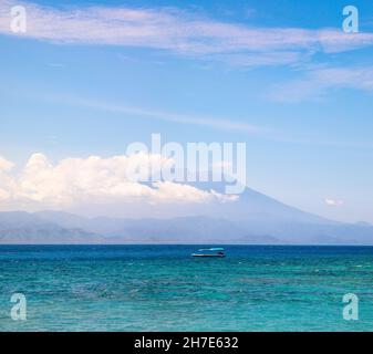 Mount Agung active volcano covered by clouds in Bali Indonesia. Traditional fishing boats called jukung on the white sand beach. High waves with foam