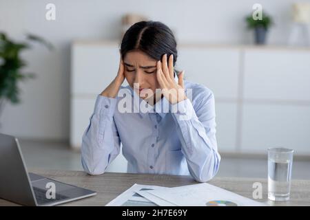 Tired upset unhappy young indian lady suffering from headache at workplace in office interior Stock Photo