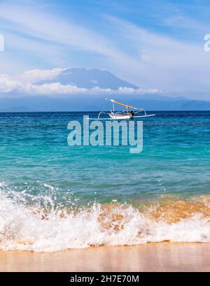 Mount Agung active volcano covered by clouds in Bali Indonesia. Traditional fishing boats called jukung on the white sand beach. High waves with foam
