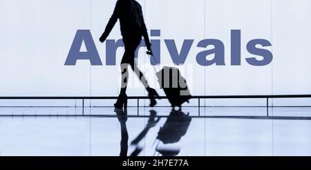Woman with wheeled luggage/suitcase passing arrivals sign in airport terminal. Stock Photo