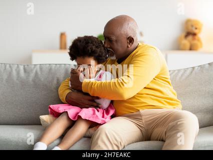 Black elderly man calming upset sad crying little girl in living room interior Stock Photo