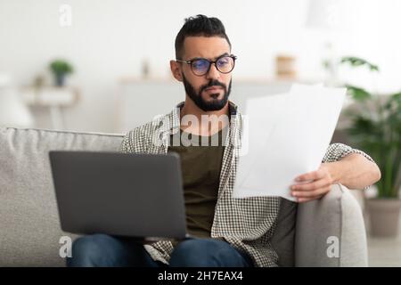 Focused young Arab guy reading documents, having online business conference at home office Stock Photo