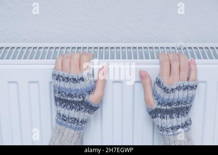 Woman in gloves warming hands on heating radiator. Winter heating capacity  Stock Photo