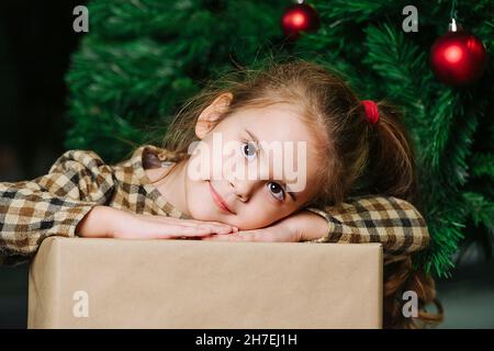 Cute girl lying her head for on her hands on a wrapped present box, posing. Sitting under a christmas tree decorated with red shiny balls. Stock Photo