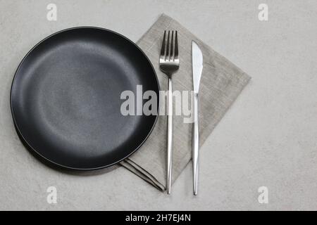 Black empty plate with knife and fork with gray textile napkin on grey stone background. Table setting. Top view.  Stock Photo