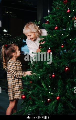 Grandma and little girl talking while decorating a christmas tree with red shiny balls. Indoors Stock Photo