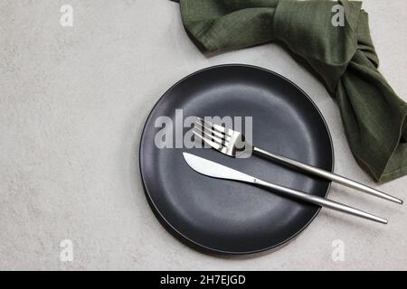 Black empty plate with knife and fork with green textile napkin on grey stone background. Table setting. Top view.  Stock Photo