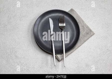 Black empty plate with knife and fork with gray textile napkin on grey stone background. Table setting. Top view.  Stock Photo