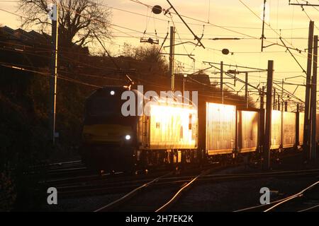 Sunset glint reflecting off electric loco and containers as freight train passes through Carnforth on West Coast Main line on 22nd November 2021. Stock Photo