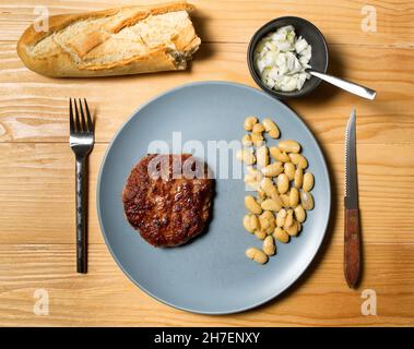 Tasty grilled burger served with coocked vegetarian beans in a plate on a rustic wooden table. Flat lay, top view Stock Photo
