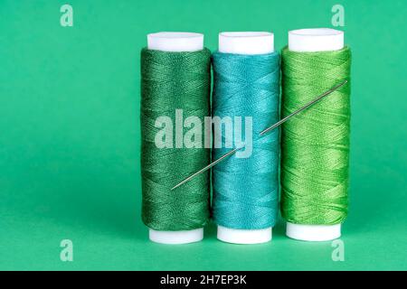 Three spools of sewing thread of different shades of green with a needle stuck in and isolated on green background Stock Photo