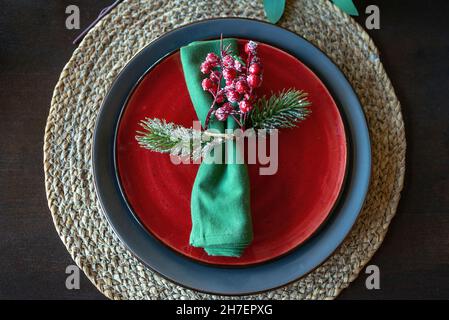 Festive christmas table setting with green napkin decorated with a sprig with berries and fir branch, red and blue plates on wooden table. Holiday Table. Top view. Stock Photo