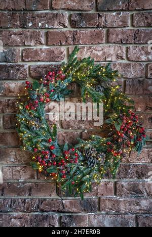 A beautiful Christmas wreath isolated on a brick wall background. Handmade Xmas wreath made from fir, red berries and pine cones hanging on wall for holiday season greeting. Vertical shot Stock Photo
