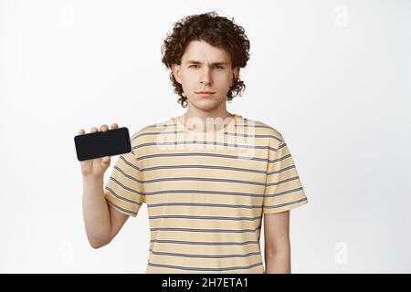 Disappointed young man showing horizontal smartphone screen, frowning and squinting, staring suspicious, standing over white background Stock Photo