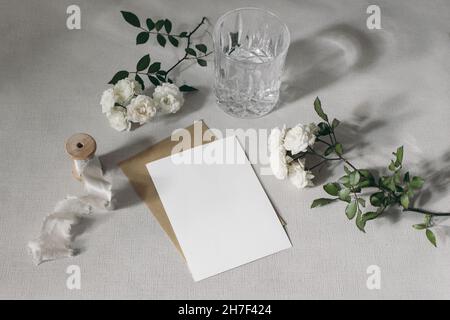 Wedding stationery mockup scene. Blank greeting card, invitation, craft paper envelope and little white roses. Sparkling glass of water on grey table Stock Photo
