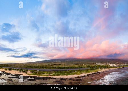 a faint rainbow forms as the setting sun lights up pink clouds over the slopes of Hualalai Volcano, as seen from Old Kona Airport County Park, Hawaii Stock Photo