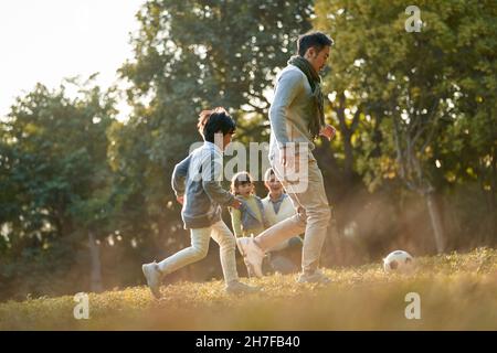 asian father and son playing soccer outdoors in park while mother and daughter watching from behind Stock Photo