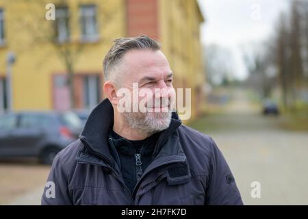 Bearded man looking aside with a quiet smile of satisfaction as he walks through a quiet urban street with colourful houses in a warm winter jacket Stock Photo