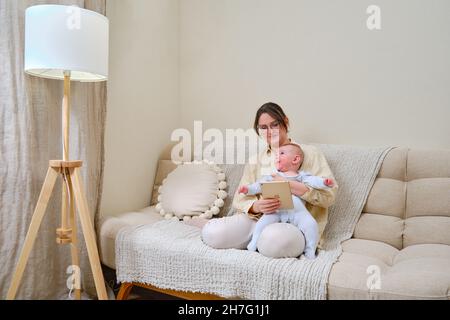 Happy woman mother with infant baby watching in a digital tablet iPad Apple while sitting on the home sofa in the living room - Moscow, Russia, Novemb Stock Photo
