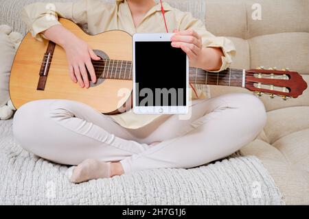 Woman musician with an acoustic guitar holds a tablet Apple iPad, mockup. Female guitarist sitting on a bed in a home living room with a tablet displa Stock Photo