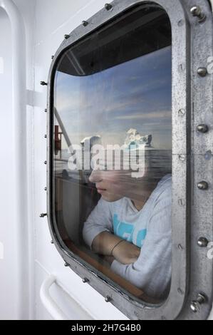 DENMARK. GREENLAND. WEST COAST. WOMAN LOOKING AT AN ICEBERG THROUGH THE WINDOW OF A BOAT. Stock Photo