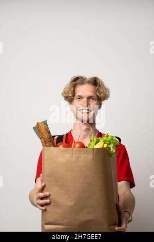 Portrait of pleased delivery man in red uniform smiling while carrying paper bag with food products isolated over white background. Stock Photo
