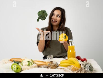 Happy woman in green t-shirt unpacking shopping mesh eco bag with healthy vegetables, fruits, bread, snacks on the kitchen at home and throwing Stock Photo
