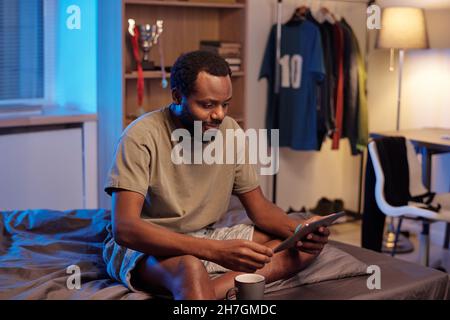 Young African man in t-shirt scrolling through news in tablet while sitting on bed after sleep in the morning Stock Photo