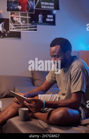 Young serious African sportsman in activewear sitting on bed and watching online game on screen of digital tablet Stock Photo