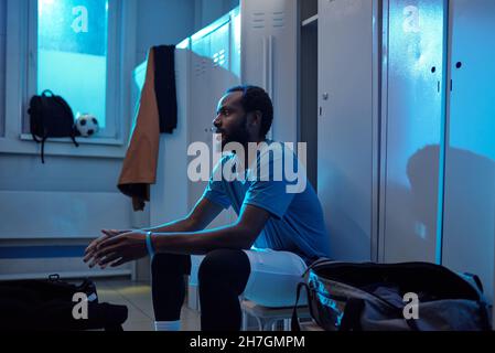 Young restful professional soccer player in sports uniform sitting against lockers in changing room Stock Photo