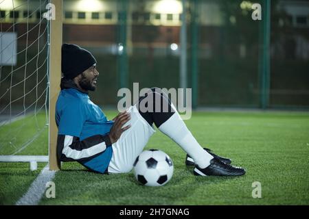 Young African footballer in professional sports uniform sitting by net on green lawn and having rest at break Stock Photo