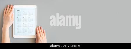 tablet computer with an open app of calendar for 2022 year in womans hands on a gray background. concept business or to do list goals with technology Stock Photo