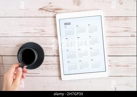 tablet computer with an open app of calendar for 2022 year and cup of tea or coffee in womans hands on wooden background. concept business or to do li Stock Photo