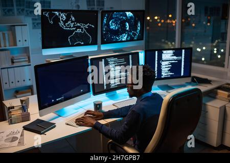 Young contemporary software developer working with decoded data while sitting in front of computer monitor in office at night Stock Photo